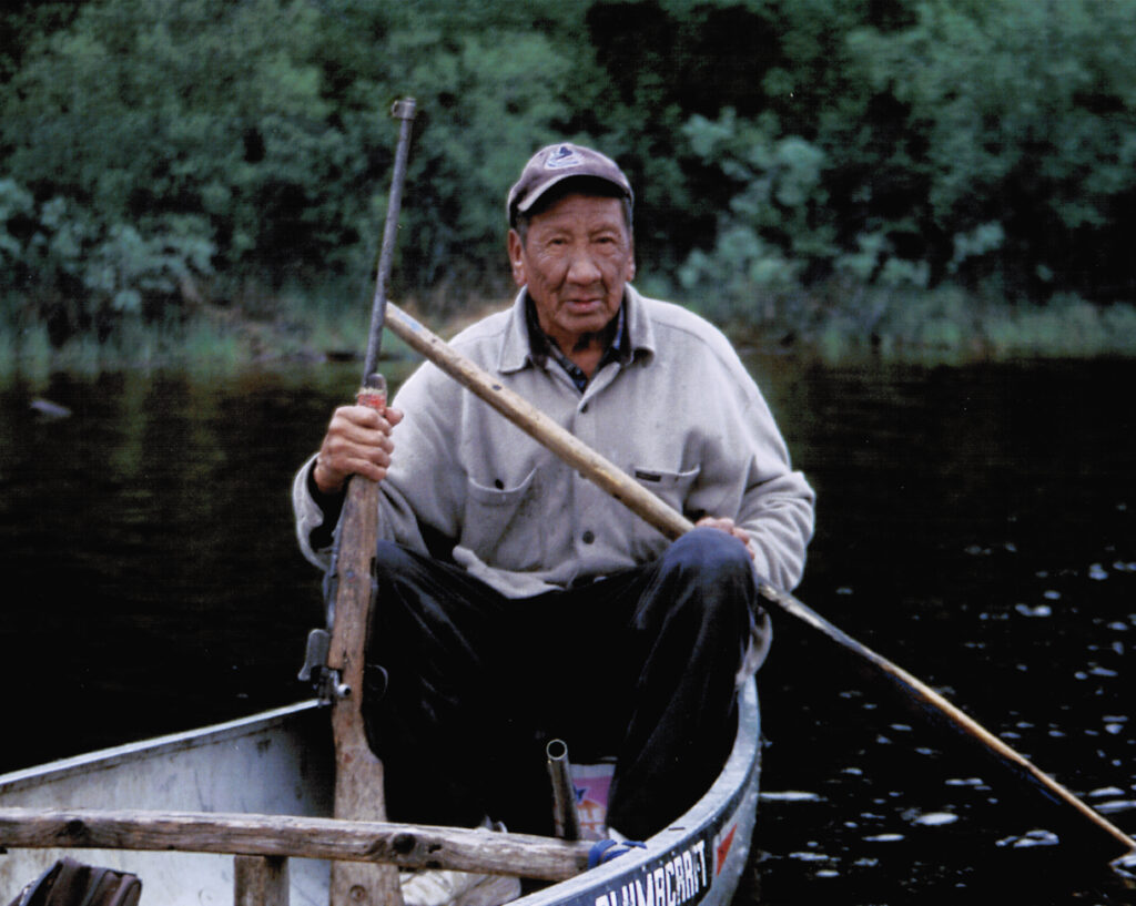 Frank Duck sits in canoe with a paddle and gun in hand.